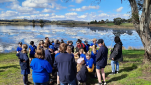 NITA Education with Oatlands primary school children at Lake Dulverton as part of the Tasmania Island Ark program. Students stand under a tall tree, overlooking a light blue river that is reflecting the white, fluffy clouds above it.