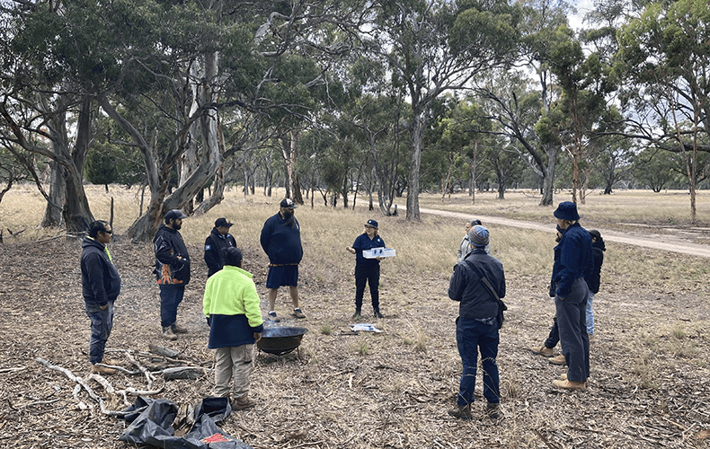 A group of people stand outside in a circle around a fire pit, talking. Gum trees are visible in the background.