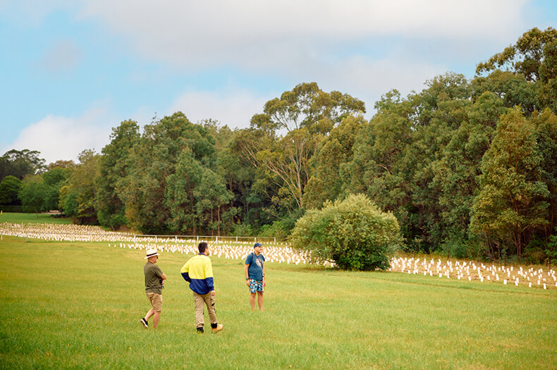 A man dressed in PPE talks to two other men who are dressed much more casually. They are standing outside on an expanse of grass and visible to one side is an extensive planting, behind which are established trees.